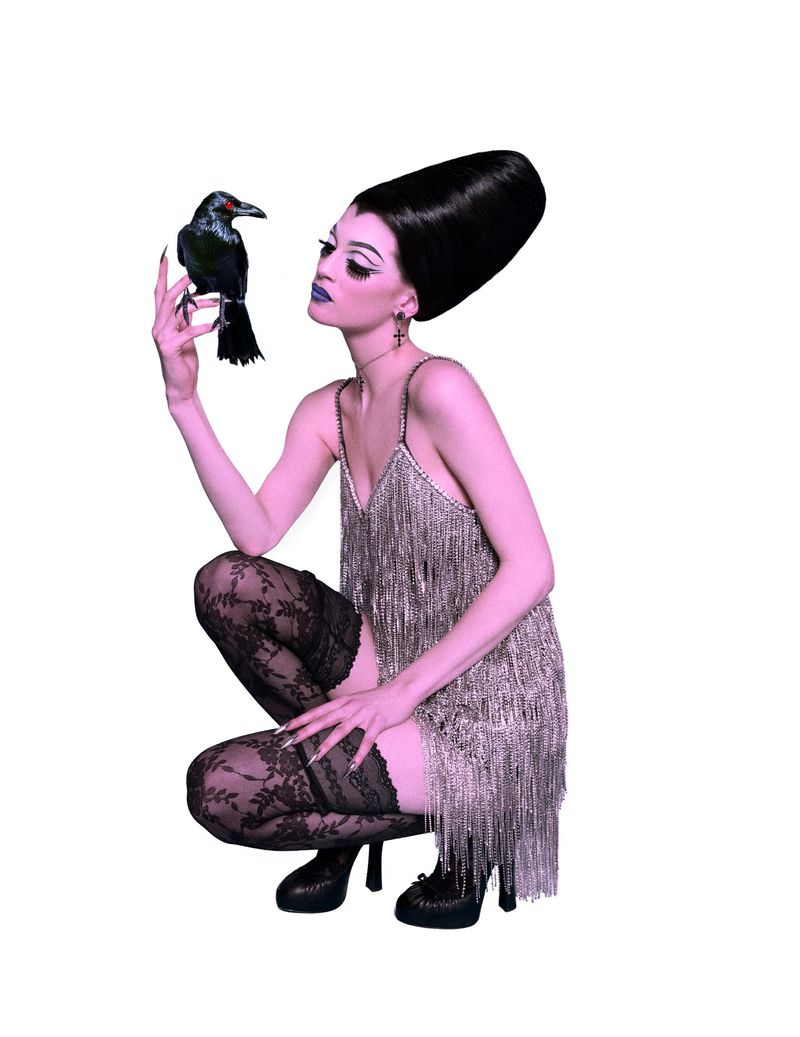 Woman in fringed dress poses with a raven against a white backdrop. She is crouching, wearing lace stockings and striking makeup.