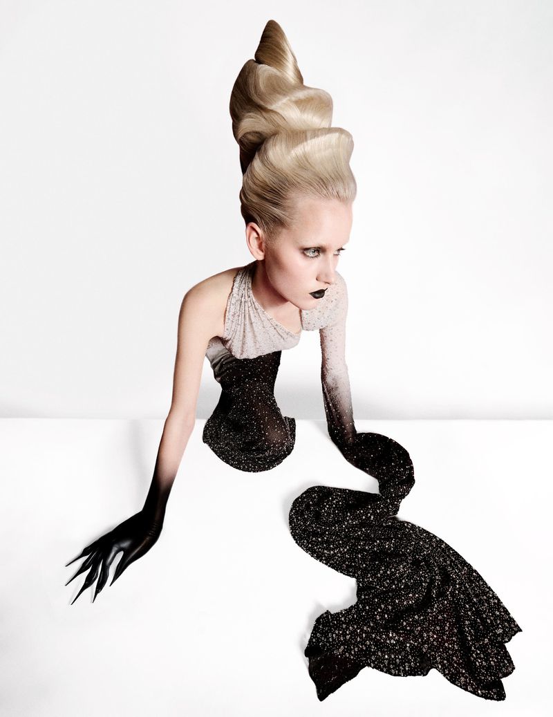 Model with extreme tall blonde hair and black lipstick poses against a white studio background with black fingernails and fashion dress.