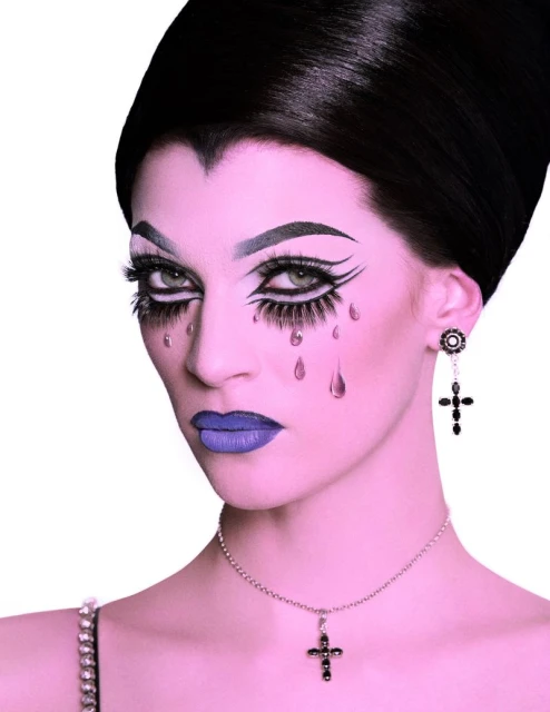 Close-up of a woman with dark, stylized makeup, dramatic eyelashes, and dark hair, wearing jewelry against a white background.
