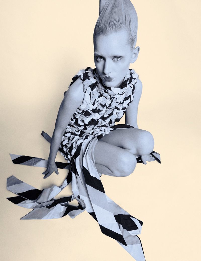 High-angle shot of a female fashion model in a black-and-white dress, crouching on a pale background, showcasing avant-garde style and design.