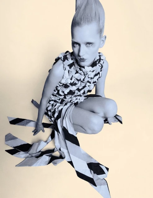 High-angle shot of a female fashion model in a black-and-white dress, crouching on a pale background, showcasing avant-garde style and design.