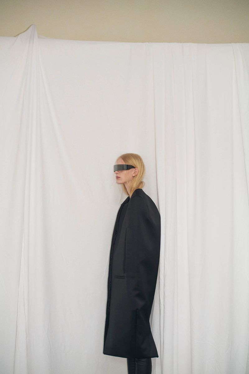 Blonde woman in long black coat and futuristic visor poses in front of a white curtain, creating a minimalist fashion editorial shot.