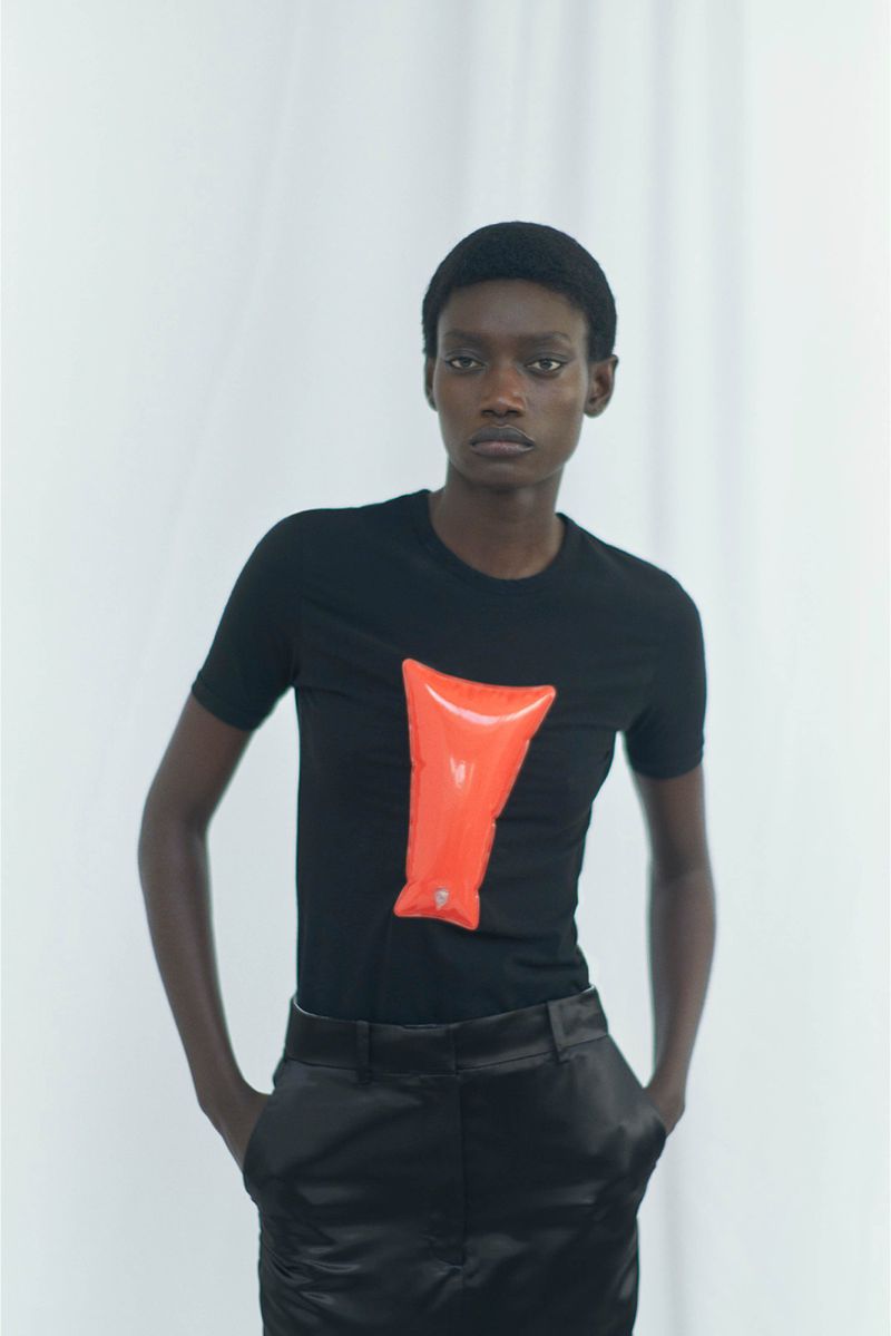 Fashion shot of a black female model wearing a black t-shirt with an orange inflatable object in front of a white backdrop.