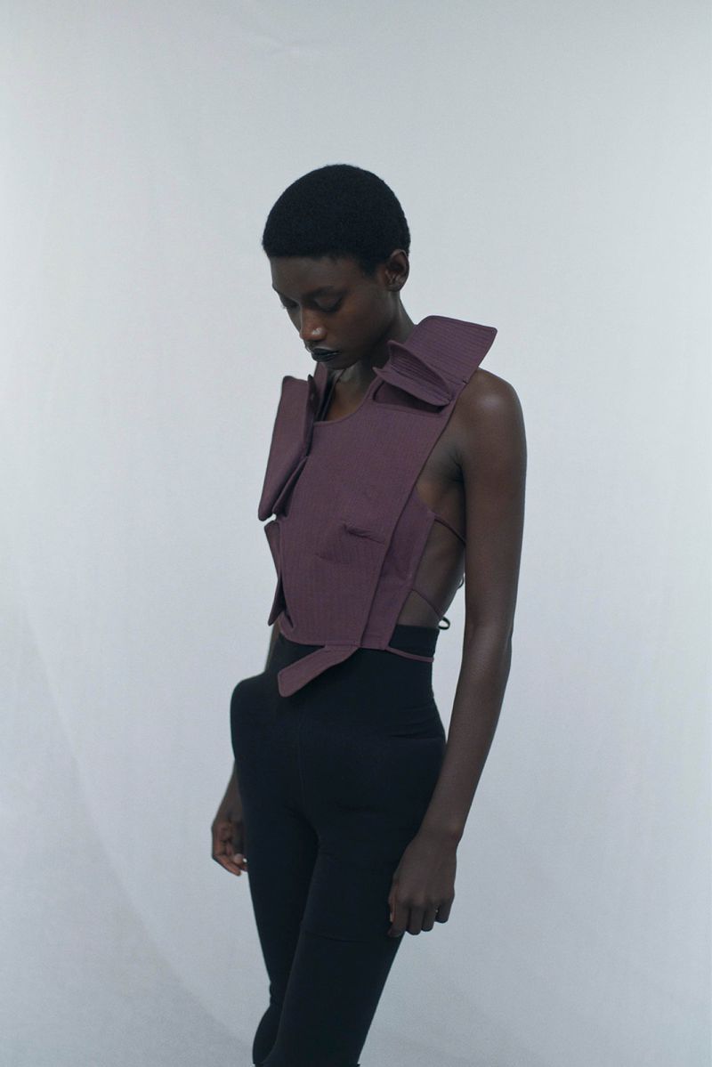 Fashion shoot featuring a single adult female model with short hair, posing in a unique purple top and black pants against a white background.