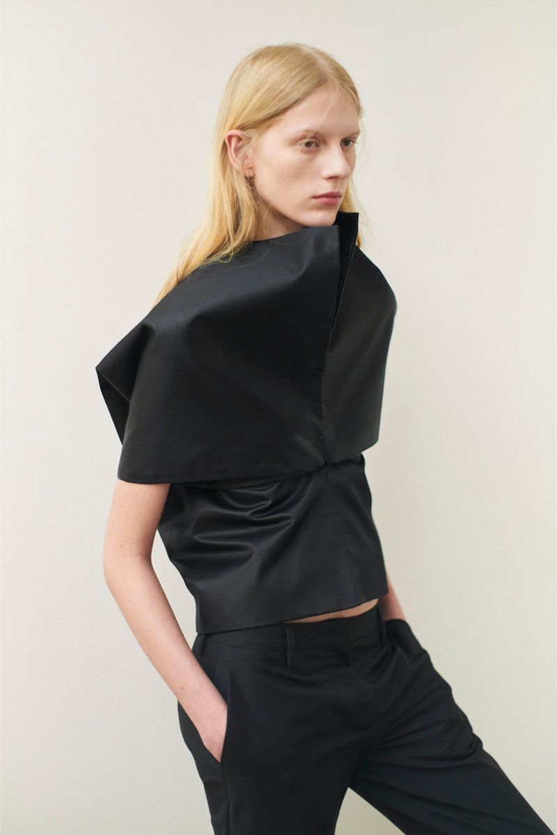 Fashionable blond model poses in a striking black satin top and matching trousers against a neutral backdrop, showcasing modern elegance.