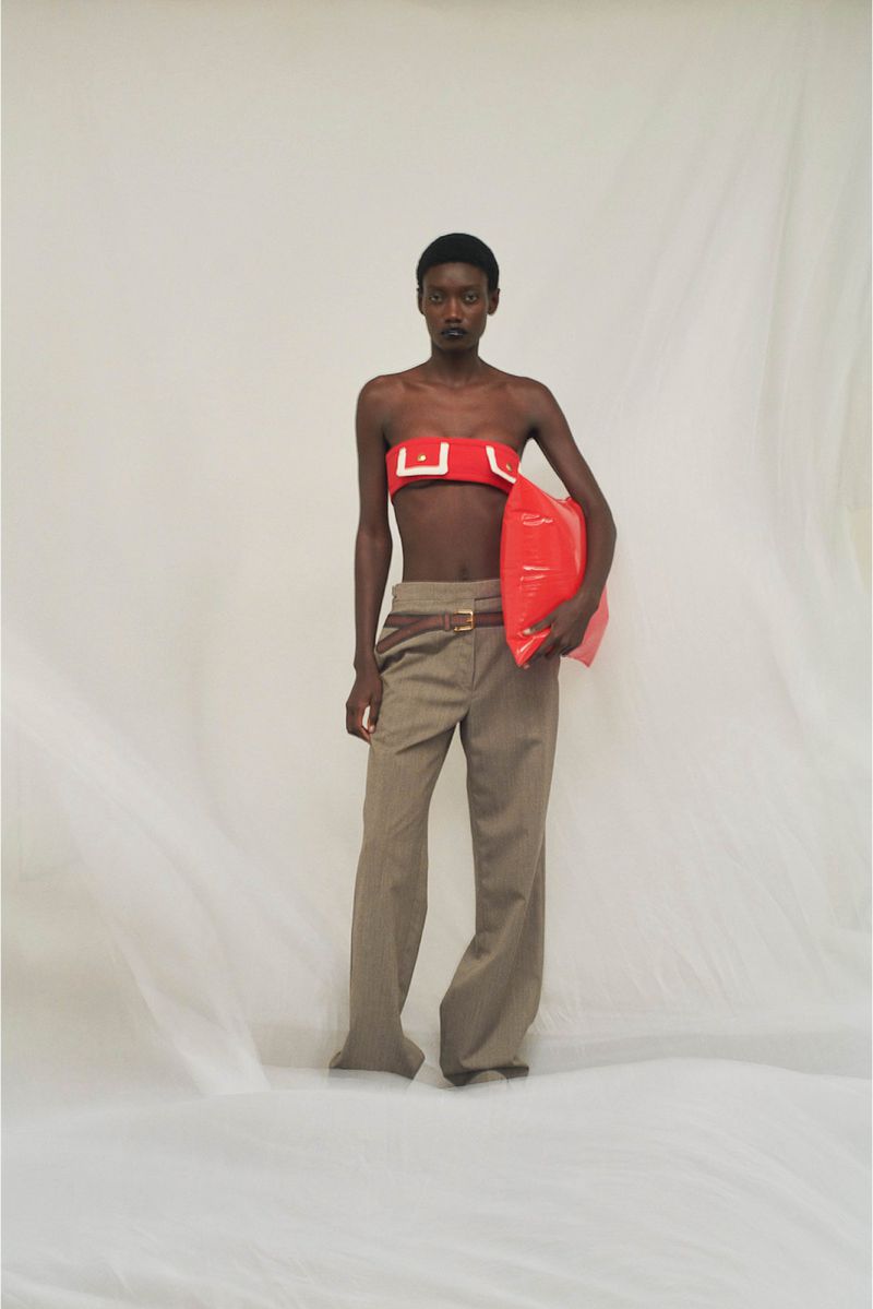 A dark-skinned fashion model poses in a studio wearing a red top and tan pants while holding a red inflatable object.