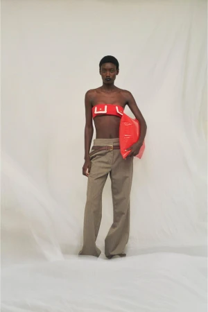 A dark-skinned fashion model poses in a studio wearing a red top and tan pants while holding a red inflatable object.