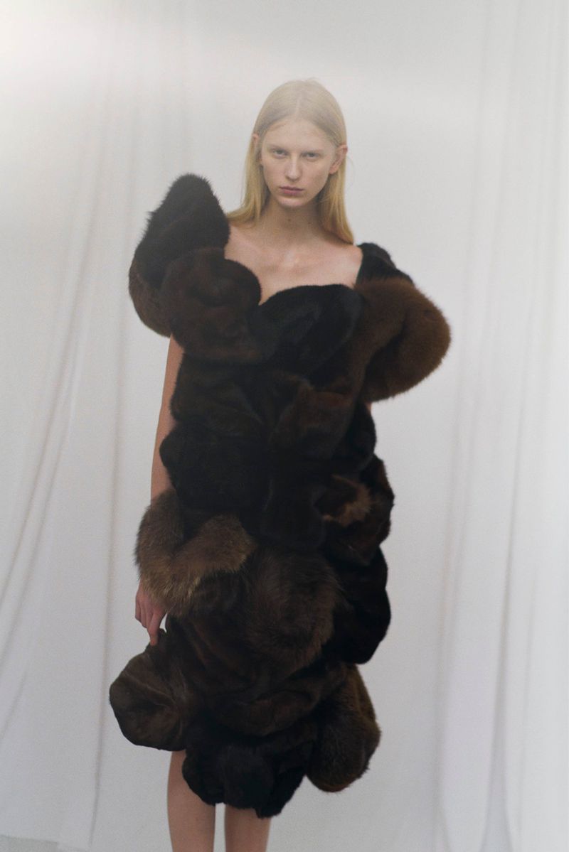 Blonde woman poses in an artful dress made of brown and black fur in a studio with soft, diffused lighting and a white backdrop.