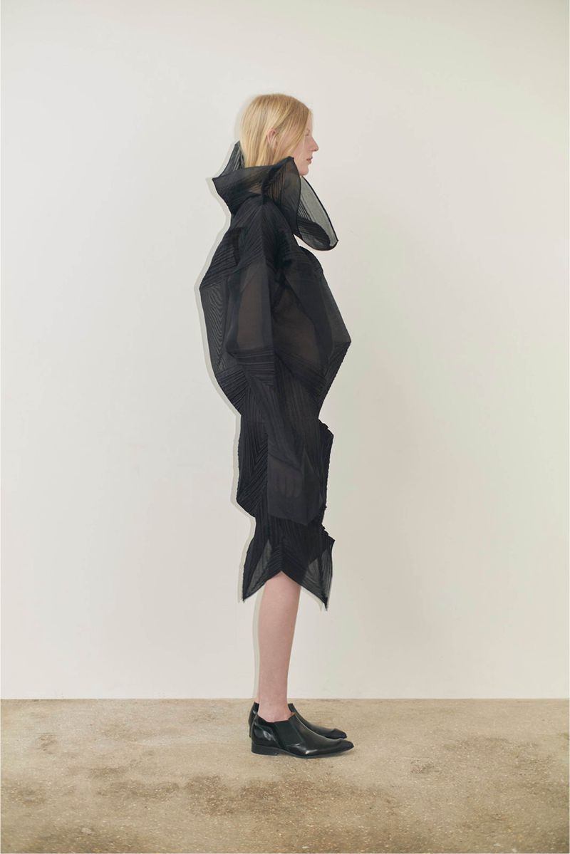 Blonde woman in a structural black dress poses in a studio with a white backdrop, showcasing fashion design and studio photography.