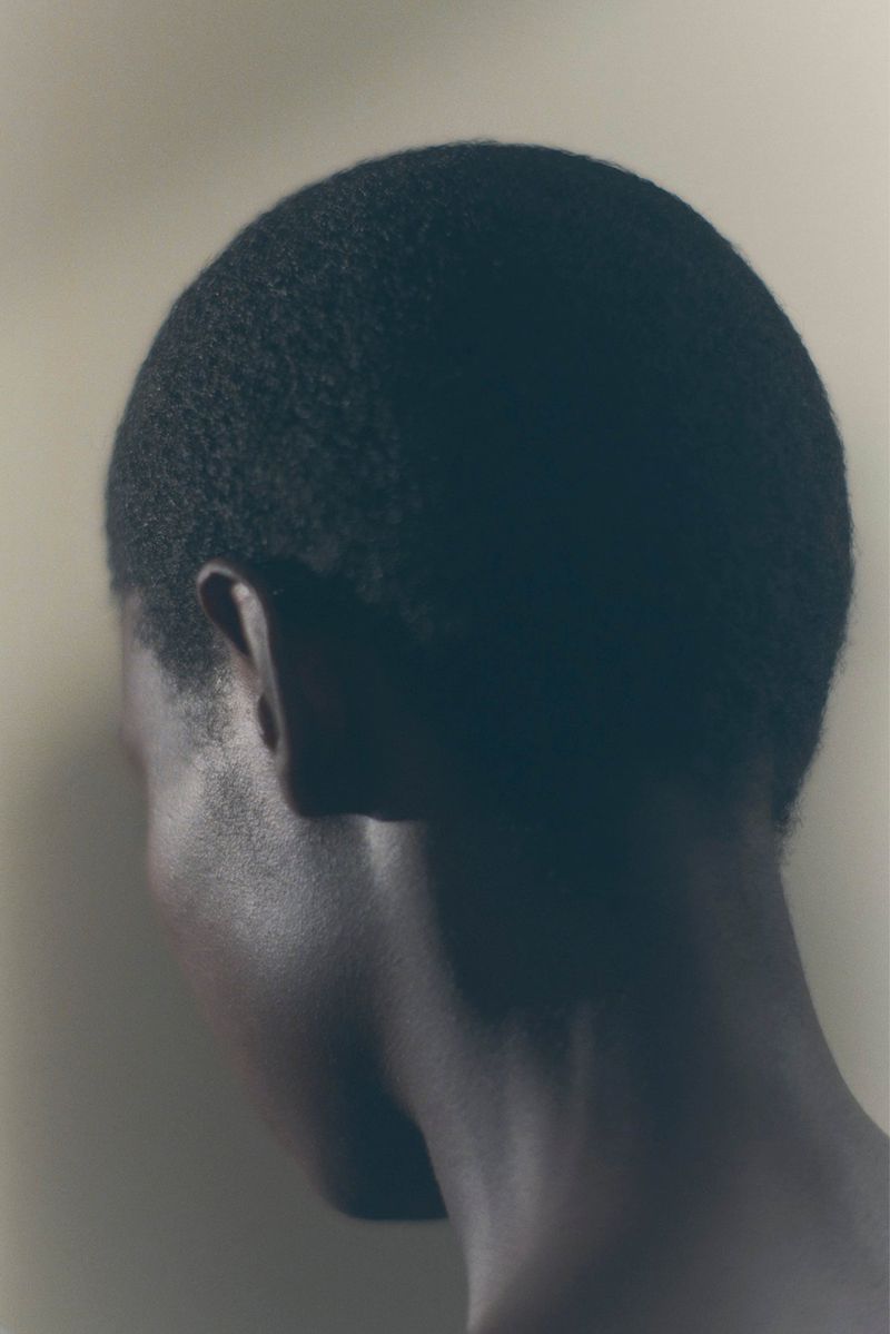 Side view of a dark-skinned adult male with closely cropped hair against a muted beige background in a portrait-oriented composition.