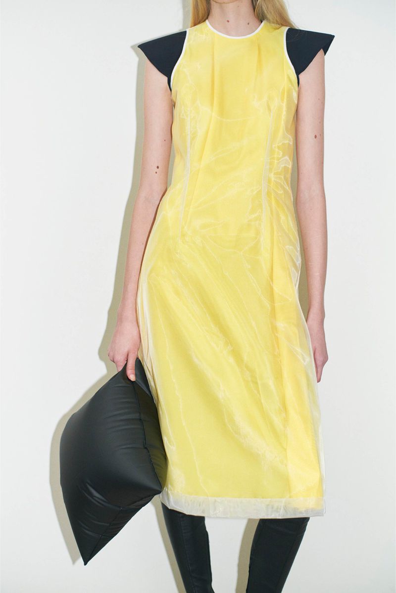 Woman poses in a yellow dress with black shoulder pads, black leggings, and black pillow. Shot in a bright white studio.