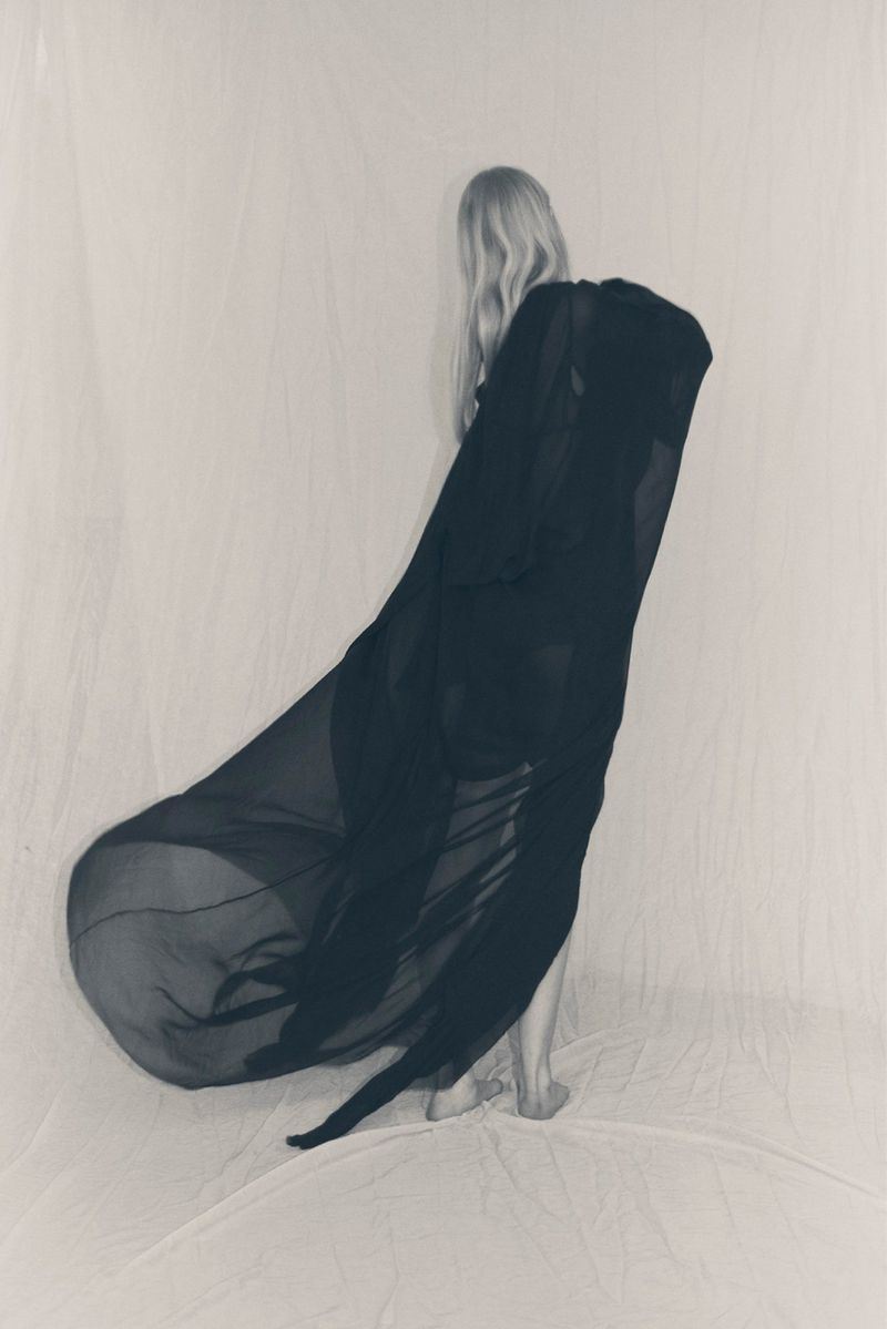 Back view of a woman with flowing blonde hair partially covered by a draped black fabric against a plain light-colored background.