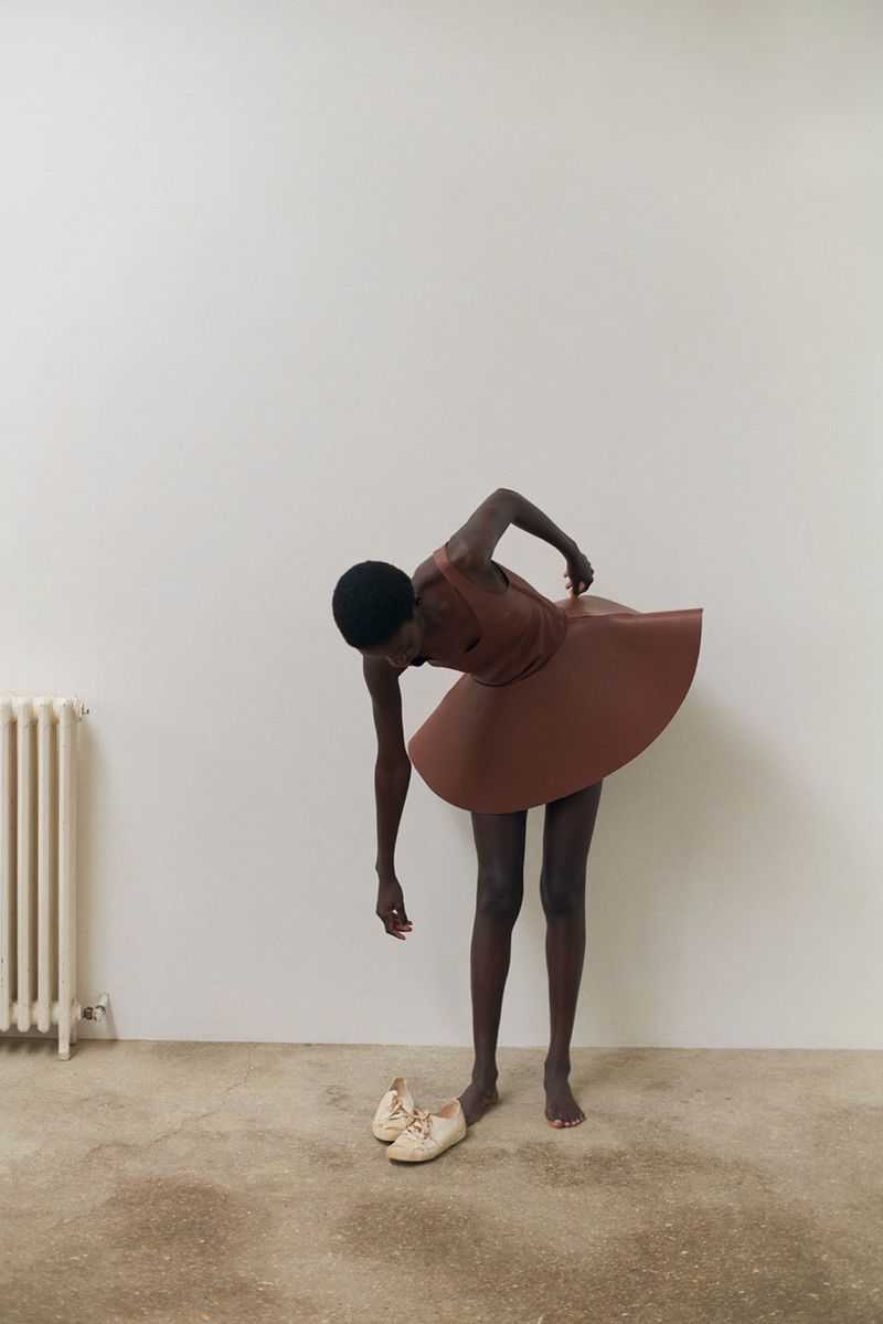 Dark-skinned person in a brown dress bending over, reaching for white shoes on a tan floor, with a white radiator in the background.