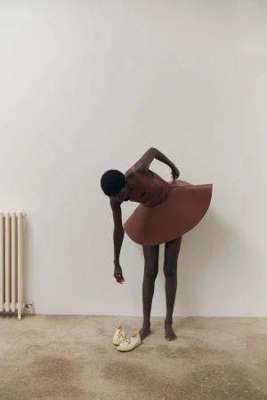 Dark-skinned person in a brown dress bending over, reaching for white shoes on a tan floor, with a white radiator in the background.