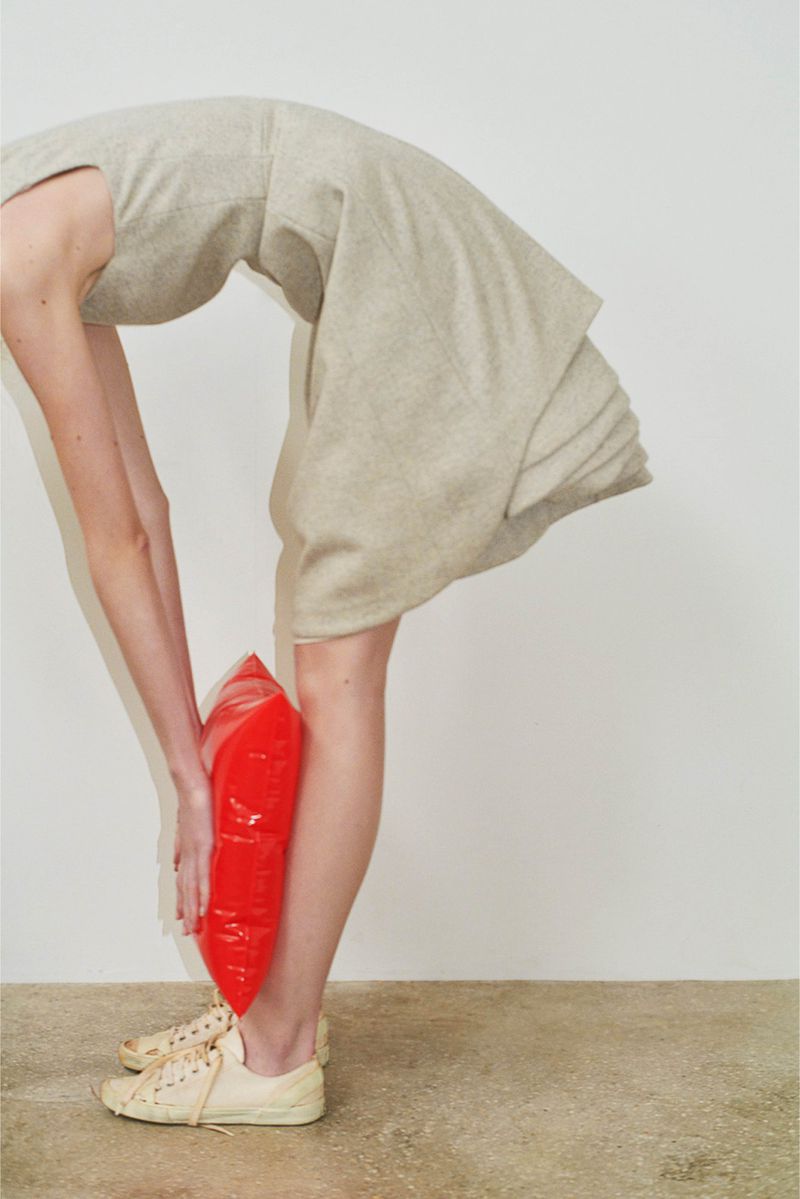 A woman in a light tan dress bends over to hold a red water balloon against her lower leg, posing in a brightly lit studio.