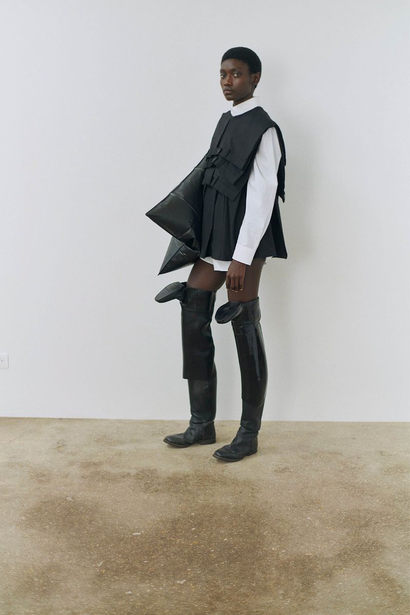 Fashion shot of a Black model in a white shirt, black vest, and thigh-high boots posing in front of a white wall in a studio.
