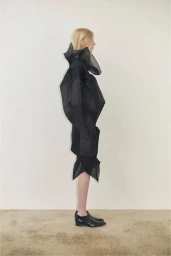 Profile view of a female model wearing a structured black sculptural garment and black shoes against a plain wall.