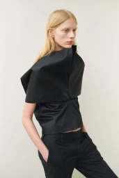 Model with long blonde hair wearing a structured black architectural top and black trousers against a neutral background
