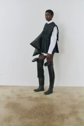 Model in black vest, white shirt, and tall boots holding a large structured bag against a plain studio background.