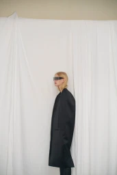 Model in profile wearing futuristic sunglasses and a dark coat against a draped white fabric background