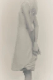 Softly blurred portrait of a woman standing against a pale seamless background wearing a simple dress.