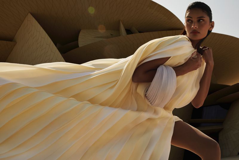 Model in cream pleated gown posing against complex modern architectural background for Vogue Arabia project Layers of Purpose