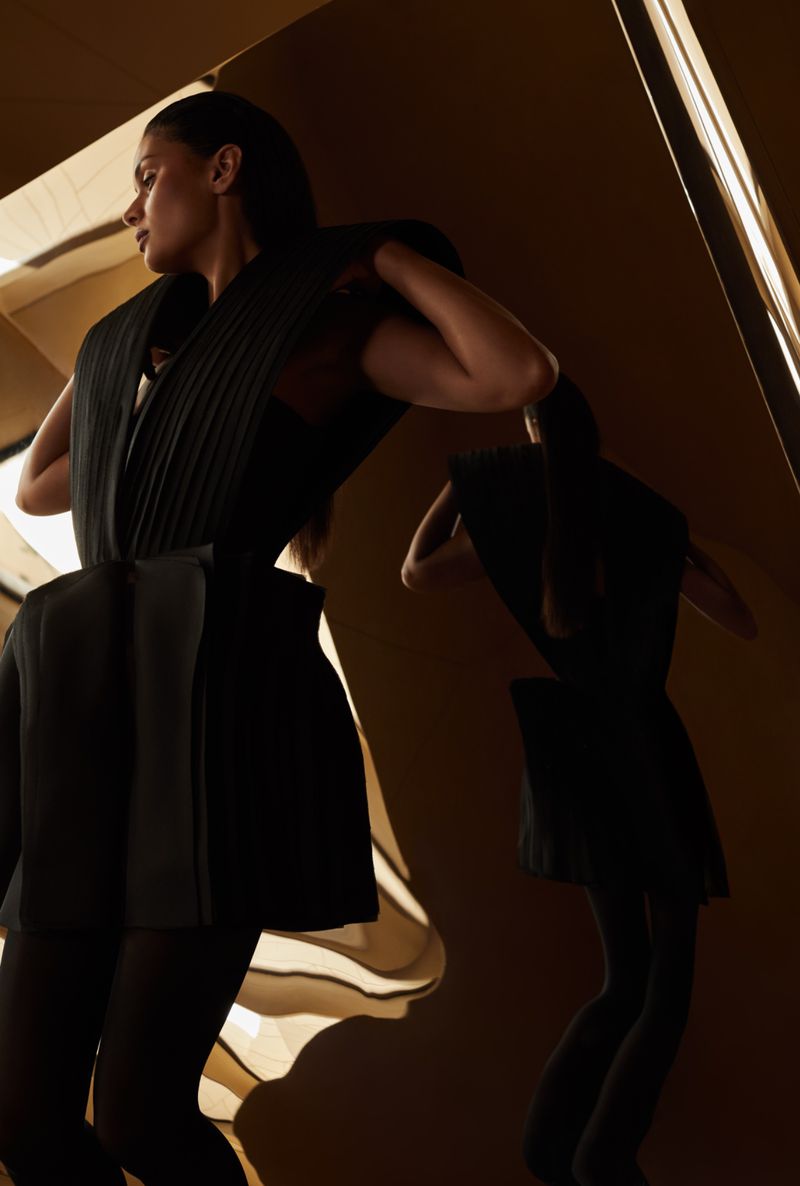 Woman in a structured black outfit poses dramatically in a studio setting with reflections and dramatic lighting in a fashion-forward shot.