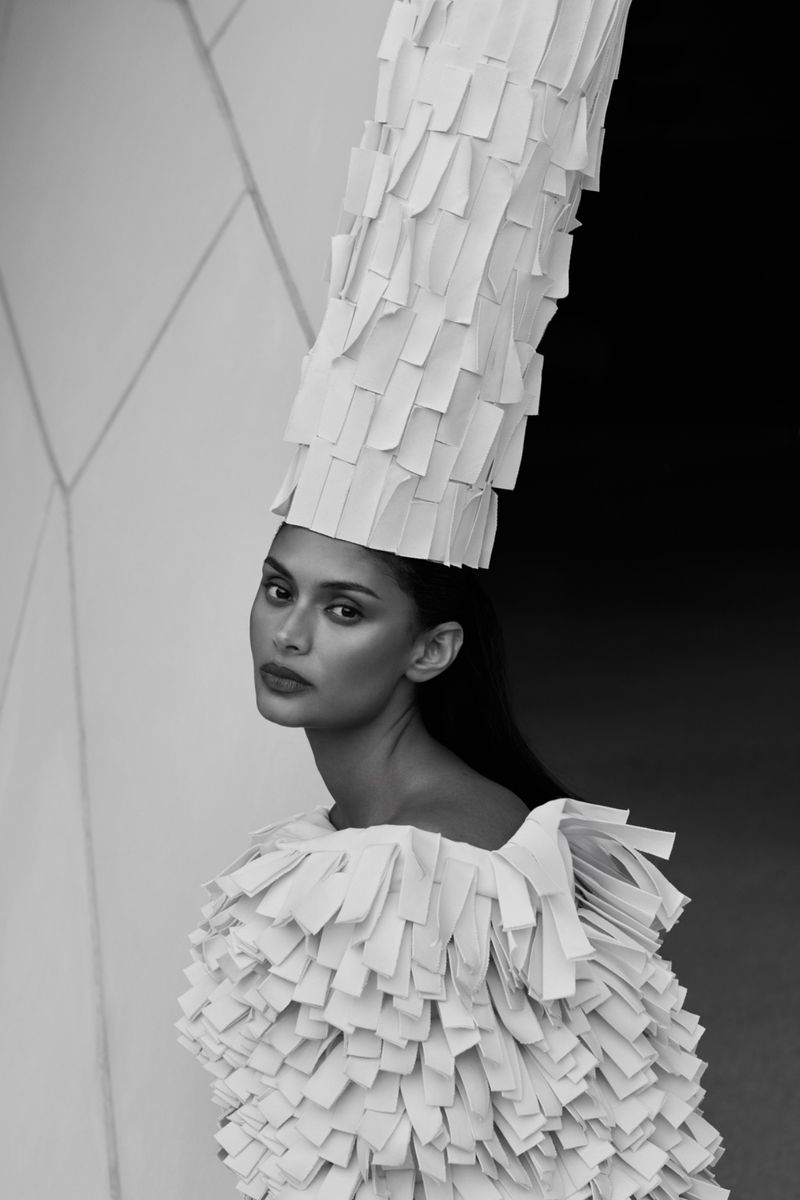 Striking monochrome portrait of a model in an artful white paper dress and tall hat, set against a textured, geometric wall.