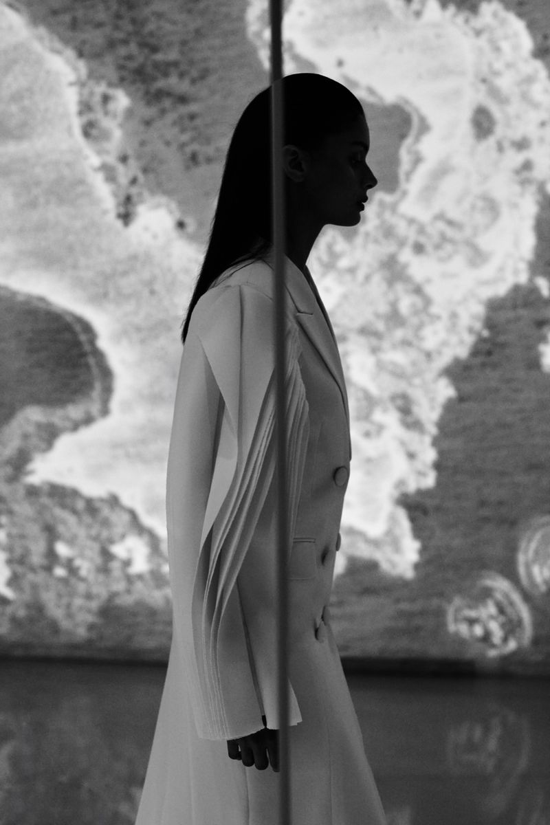 Profile of a woman with long dark hair wearing a white suit jacket, posing in front of a marble wall, black and white photography.