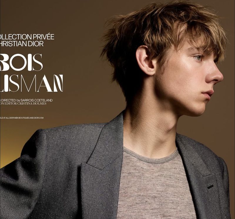 Portrait of a young blonde male model wearing a grey blazer and t-shirt, posing in a photo studio with soft lighting and golden background.