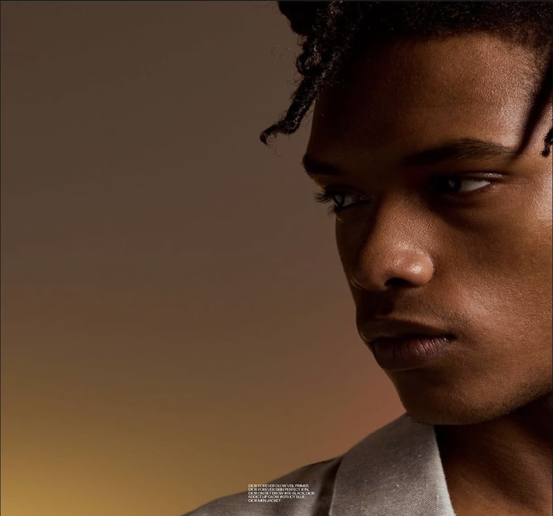 Close-up portrait of a young Black man with styled hair looking sideways against a warm gradient background.
