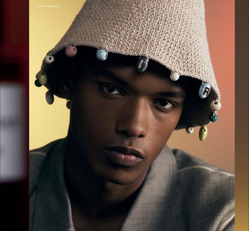 Close-up of a dark-skinned man with a serious expression wearing a tan crocheted bucket hat decorated with beads. Fashion shot.