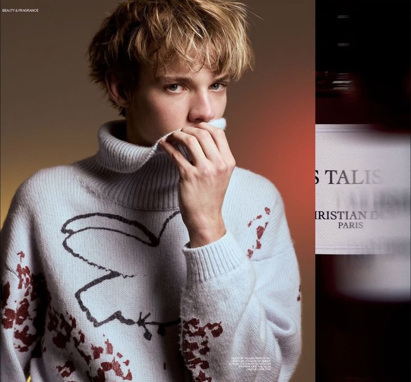 Portrait of a blond male model wearing a graphic print turtleneck sweater, with a blurred perfume bottle in the background.
