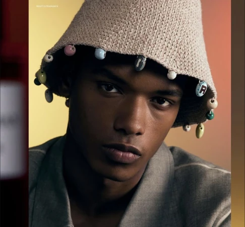 Close-up portrait of a young black man wearing a beige bucket hat adorned with colourful beads and a grey shirt against a warm gradient background.
