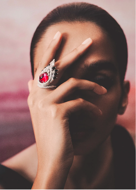 Close-up of a woman's hand with a large ruby ring, set against a soft pink background. The focus is on the jewelry and hand.
