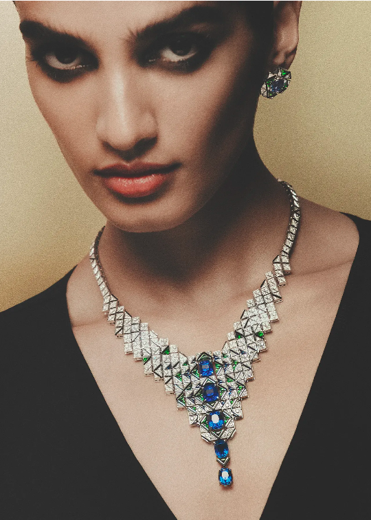 Close-up of an adult female model wearing a geometric diamond and sapphire necklace and earrings against a gold background in a studio.