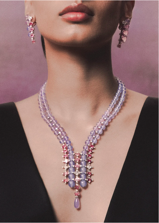 Close-up studio shot of an adult woman showcasing a lavender beaded necklace and matching floral dangling earrings against pink backdrop.