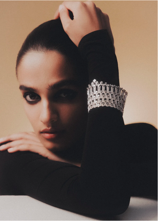 Close-up portrait of a woman wearing a wide diamond cuff bracelet on a black long-sleeve top against a warm beige background.