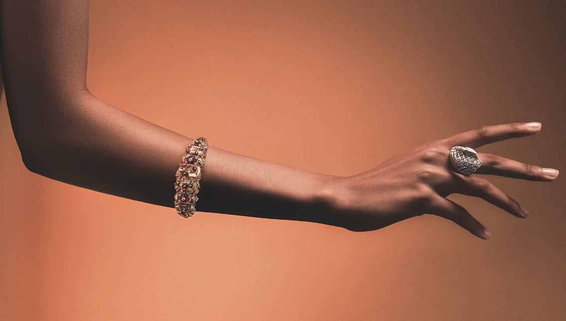 Close-up of a person wearing an ornate jeweled bracelet and a large sculptural ring against a warm brown background