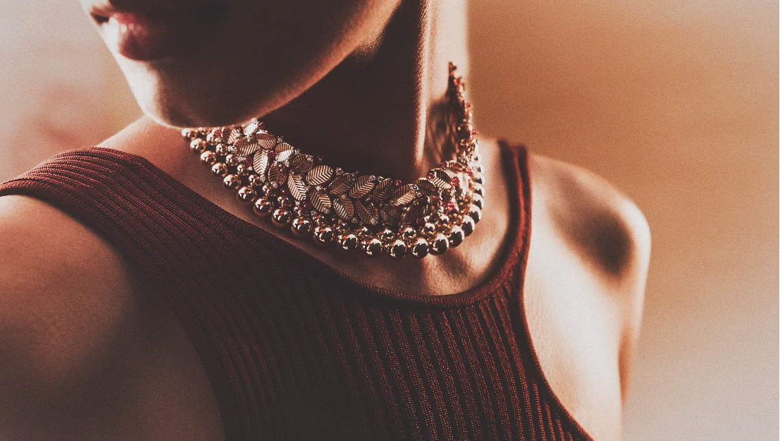 Close-up shot of a woman wearing a gold leaf statement necklace and a red tank top, bathed in warm lighting against a neutral background.