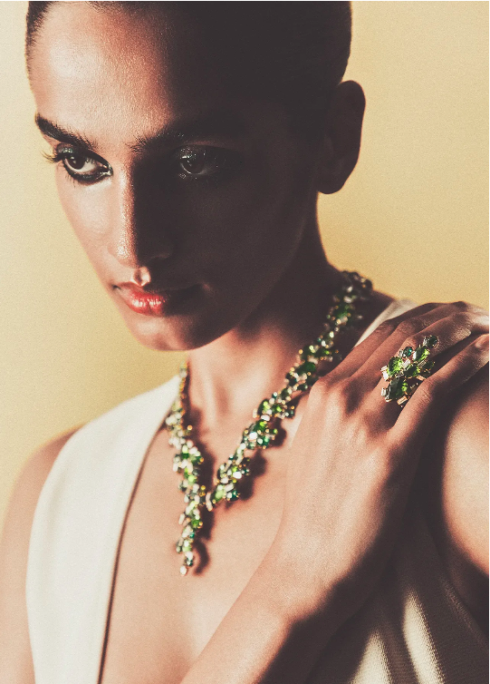Elegant woman wearing a white dress and jewelry, including a green and silver necklace and ring, against a yellow background.