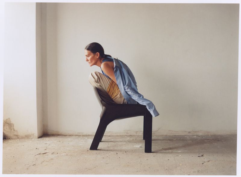 An adult woman sits on a modern chair indoors against a white wall, with natural light illuminating the scene. She's wearing beige pants and a blue shirt.