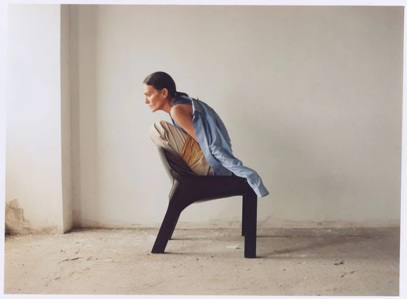 An adult woman sits on a modern chair indoors against a white wall, with natural light illuminating the scene. She's wearing beige pants and a blue shirt.