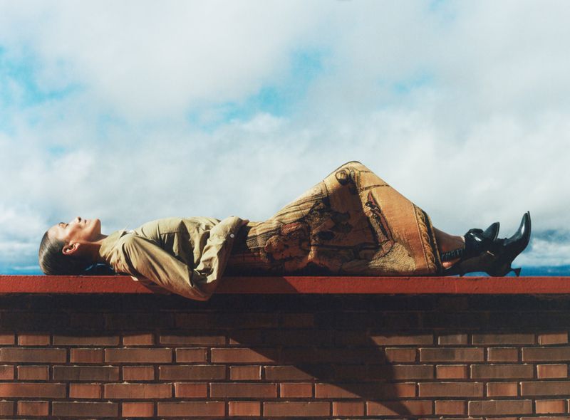 Person lying on brick ledge wearing earth-toned clothing with a blue and cloudy sky. Fashion shot, stylish shoes, outdoor setting.