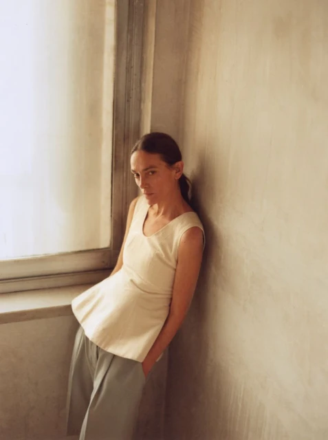 Woman posing indoors in a cream sleeveless top and blue pants, leaning against a neutral wall near a window in diffused light.