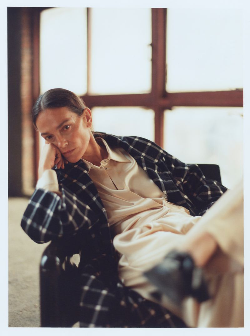Model Laura Ponte seated indoors wearing a plaid coat and pale shirt for Vogue Espana