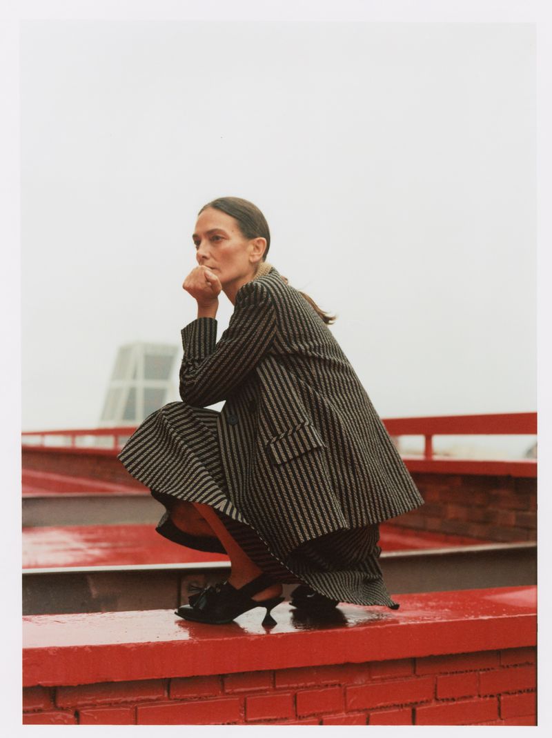 A woman in a striped blazer and skirt crouches on a red brick surface, with a modern building visible in the background on an overcast day.