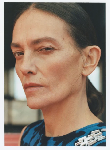 Headshot of a middle-aged woman with dark hair, wearing a blue and black patterned dress with jeweled embellishments, looking at the camera.