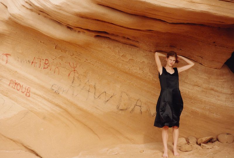 A woman in a black dress poses barefoot inside a desert cave with sandy walls featuring desert landscape and rock formations.