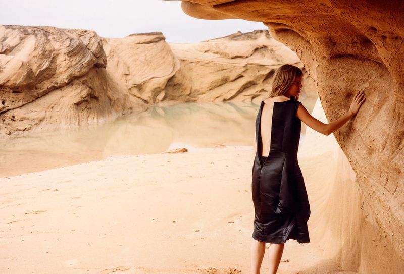 Woman in black satin dress poses near rocky landscape with water, creating a striking fashion and nature-themed visual narrative.